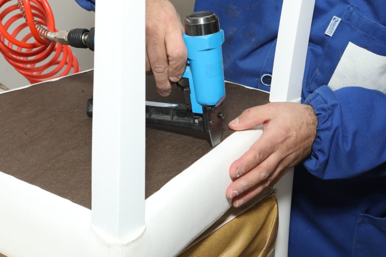 A maker upholstering a chair seat with a pneumatic staple gun, fitting cream leatherette over the frame