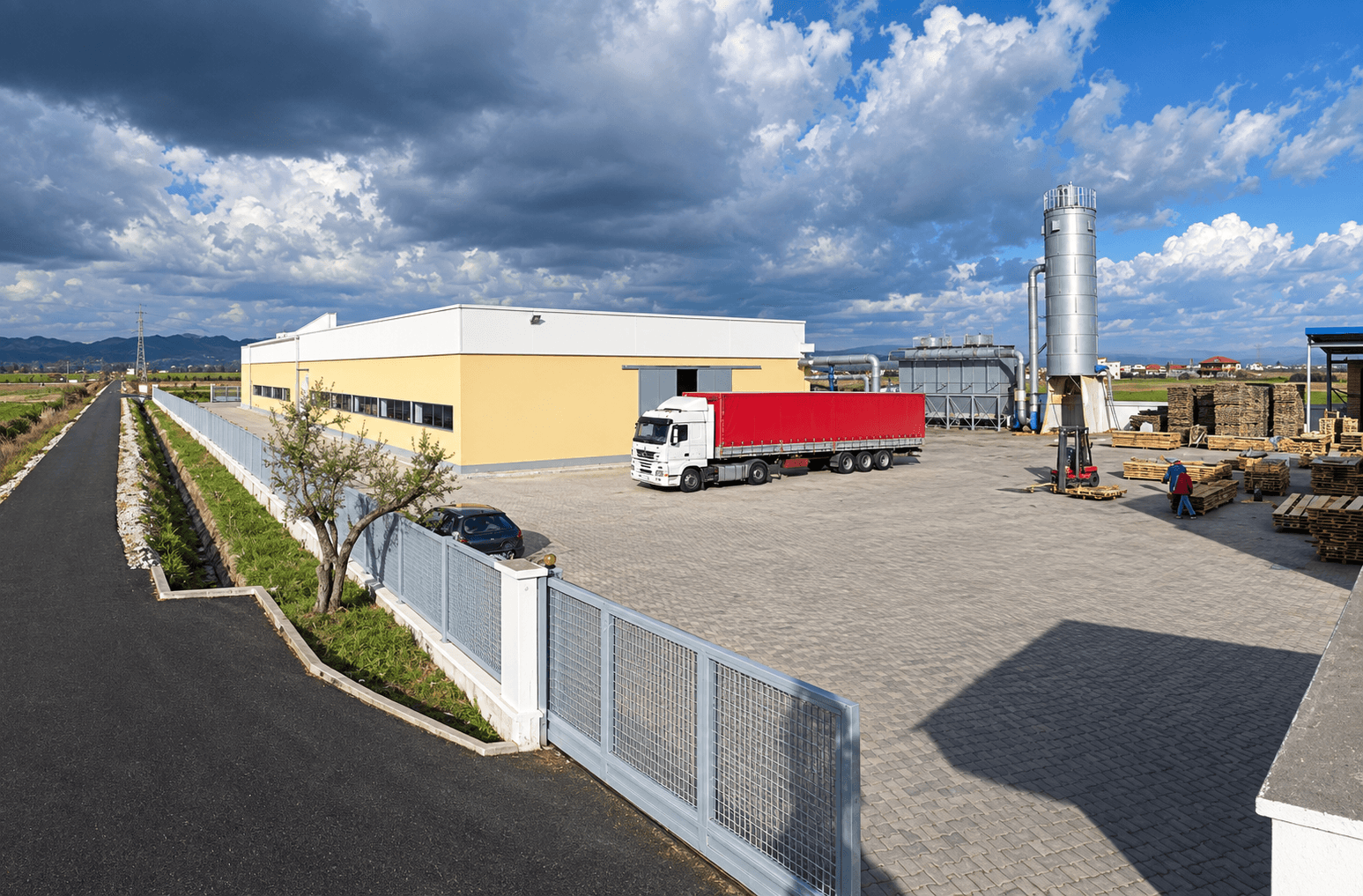 Edi Sedia factory yard with a delivery truck, storage silo and stacked pallets, mountains in the distance
